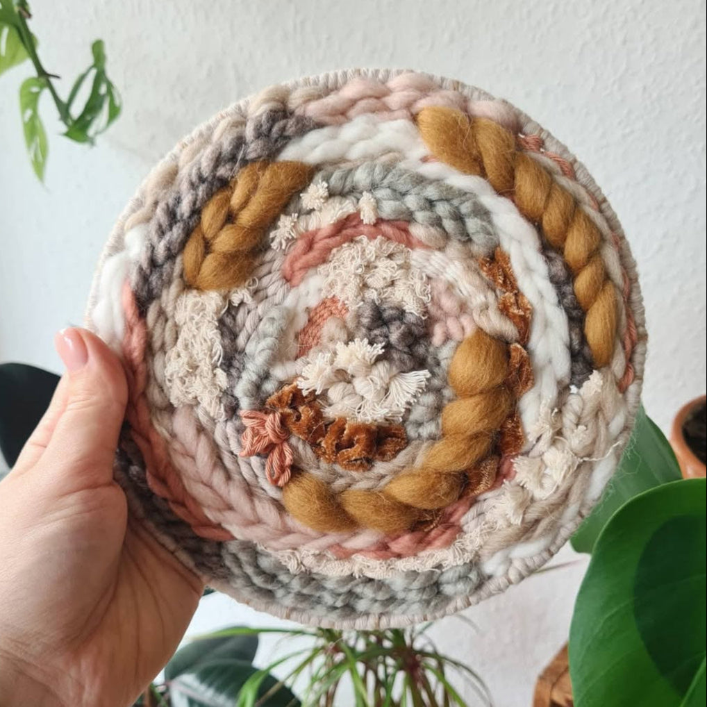 Hand holding a woven circular mat with braided edges, surrounded by plants.