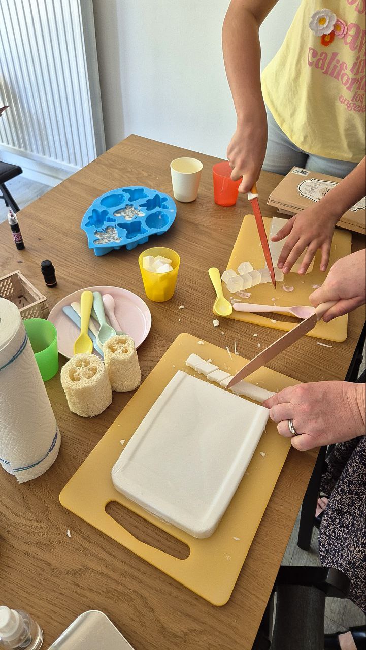Person cutting a block of white material on a wooden table with various items around.